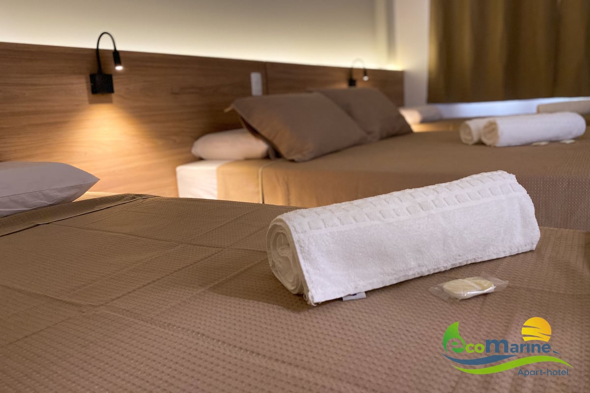 Ein Hotelzimmer mit einem ordentlich bezogenen Bett, einem Kopfteil mit Wandleuchten und dem sichtbaren Logo des 'EcoMarine Apart-hotels'.