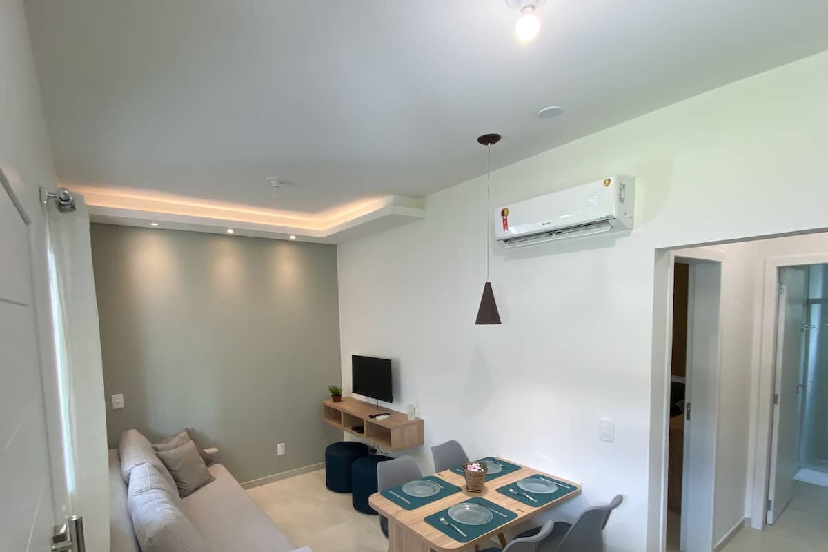 Ein modernes Wohnzimmer mit einer Couch, einem Esstisch gedeckt mit Tellern und Besteck, einem Fernseher auf einem Regal und einer Klimaanlage an der Wand.