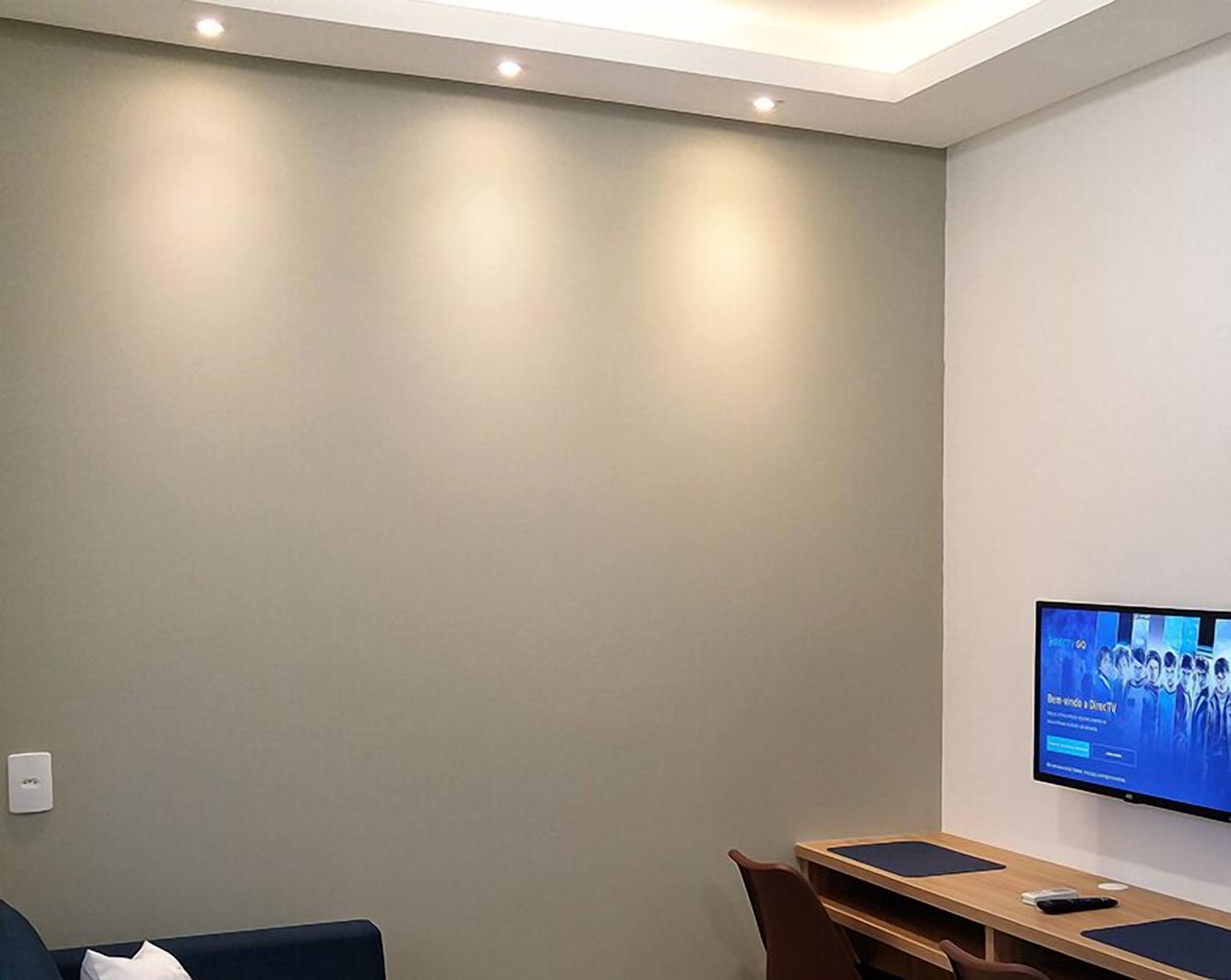 Habitación moderna con televisión de pantalla plana en la pared mostrando un noticiero. Hay un escritorio de madera con una computadora y una silla frente a él.