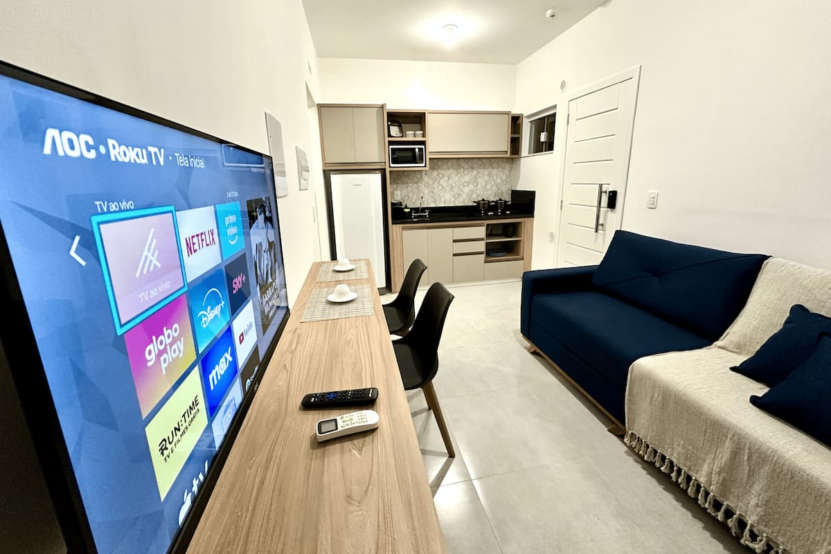 Ein modernes Wohnzimmer mit Fernseher, der Streaming-Dienste anzeigt, einem Esstisch mit Stühlen, einem Sofa und einer Küche im Hintergrund.