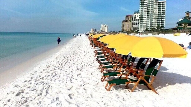 A beach with rows of beach chairs and umbrellas, a sandy shore, and buildings in the background. - Beach,Tons of entertainment, bikes, 2  golf carts, Golf, Amazing Gym, and Marina (Miramar Beach)