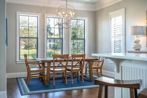 A dining area with a round table and six wooden chairs, large windows with blinds, a chandelier, and a lamp on a white console table.