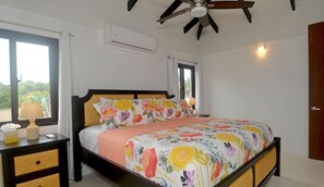 A bedroom with a bed, nightstands, an air conditioning unit, a ceiling fan, and a view of greenery outside.
