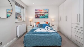 Une chambre à coucher comprenant un lit, deux tables de chevet, une grande fenêtre, un radiateur, un miroir fixé au mur, un tableau et une armoire.