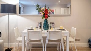 Un coin repas avec une table dressée pour quatre personnes, des chaises blanches, un vase bleu rempli de fleurs et une lampe de sol moderne.