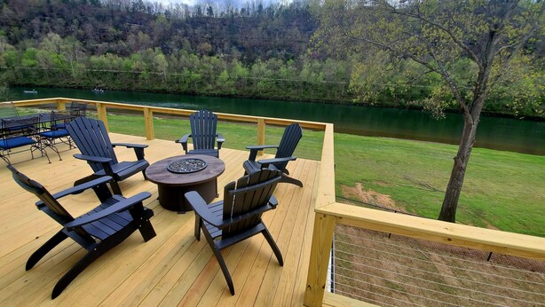 A wooden deck with outdoor furniture, including chairs and a round table, overlooking a river and forested hillside.