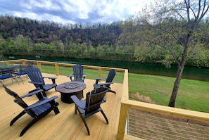 Un terrazza in legno con mobili da esterno, tra cui sedie e un tavolo rotondo, affacciata su un fiume e un pendio boscoso.