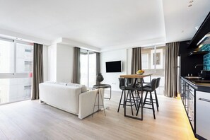 Una sala moderna con un sofá, una mesa de comedor con sillas, un televisor de pantalla plana montado en la pared y una zona de cocina con encimera y electrodomésticos.