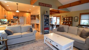 A spacious living area with a sofa, coffee table, and a bookshelf. A kitchen is visible in the background, and a sign reads "Beach House." - Cozy, year round beach cottage just steps to the water (East Haven)