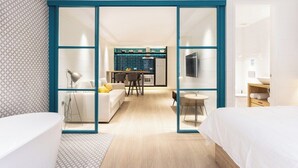 A modern hotel room with a large bed, a sitting area with a sofa and chairs, a kitchen area visible through a glass partition, and a bathroom with a bathtub.