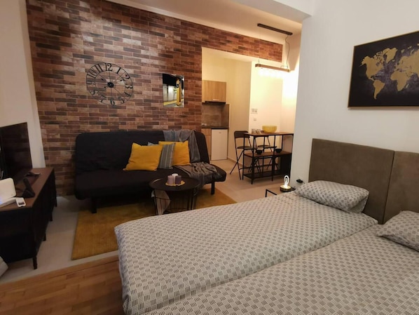 A compact hotel room with a brick accent wall, a large bed, a sofa, a small table, and a kitchenette area.