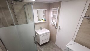 A compact bathroom with a glass shower enclosure, a white sink with a mirror, a towel rack, and a washing machine. - Charming 1-bed Apartment Located in Athina (Athens)