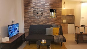 A living room with a brick wall, a round clock, a TV, a sofa, a small table, and a kitchen in the background. - Charming 1-bed Apartment Located in Athina (Athens)