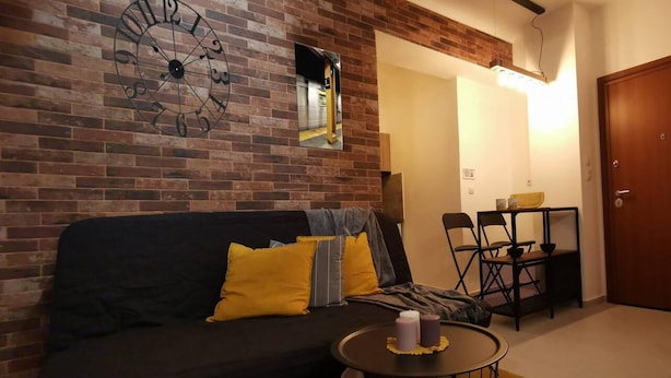 A living room with a brick wall, a black sofa with yellow cushions, a round coffee table, and a dining area with a small table and chairs. - Charming 1-bed Apartment Located in Athina (Athens)