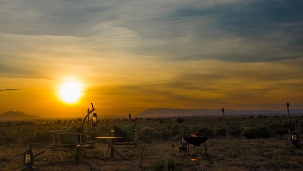 Paisaje desértico al atardecer con una fogata, una silla y una mesa lista para comer al aire libre.