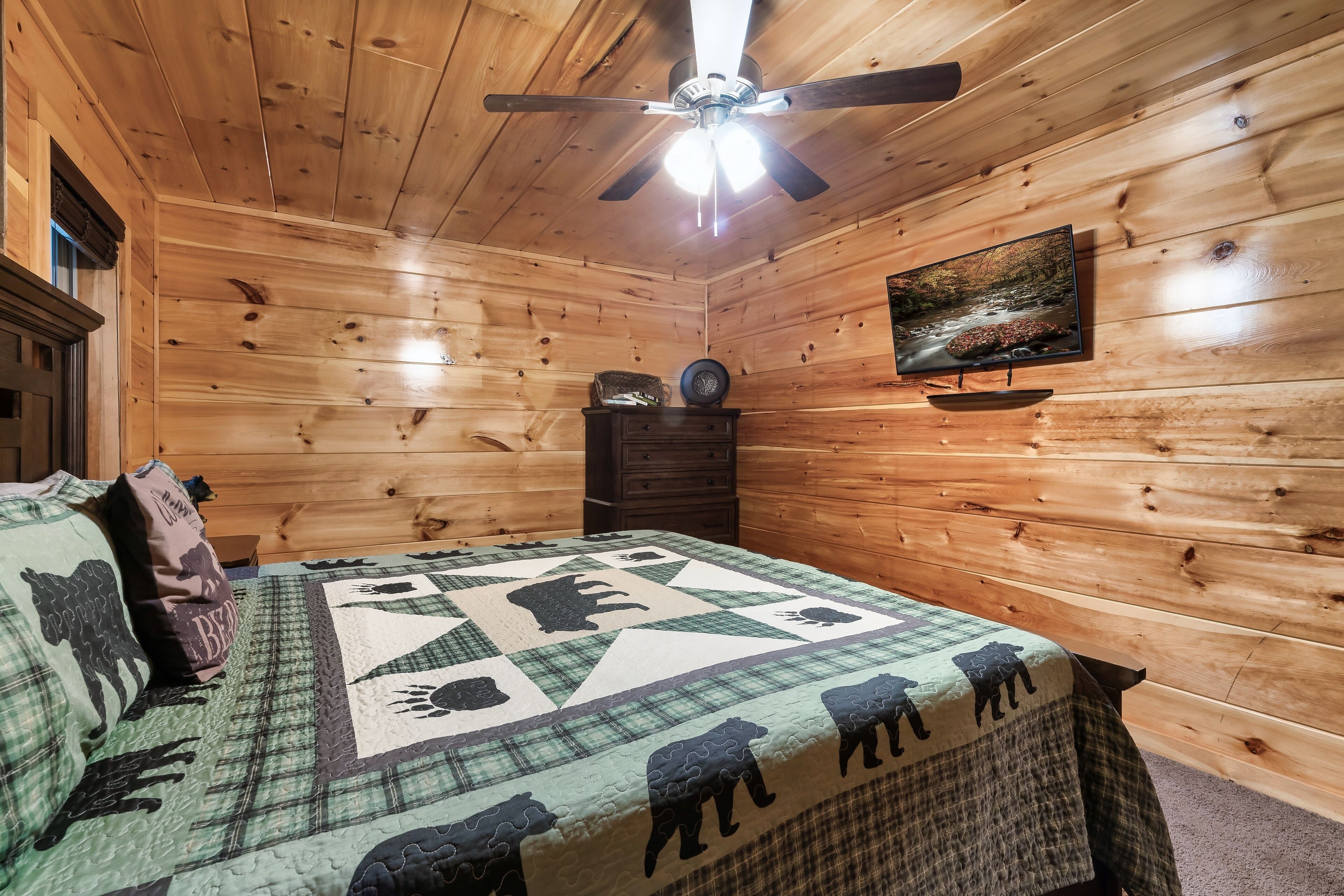 A wooden cabin room with a bed, a ceiling fan, a television, and a painting on the wall.