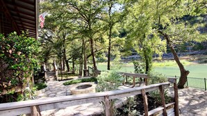 Un sendero de piedra conduce a una terraza de madera con vistas a un río, con una bandera a la izquierda y una valla metálica a la derecha.