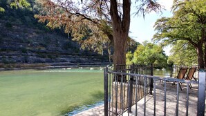 Una terraza a orillas del río con barandillas de metal y sillas, con vistas a un río tranquilo y una ladera rocosa al fondo.