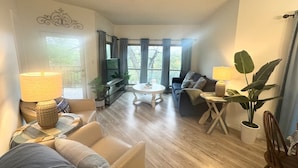 Una sala de estar con sofá, sillones, una mesa de centro y una mesa de comedor. Hay plantas en macetas y vistas al exterior a través de amplias ventanas.