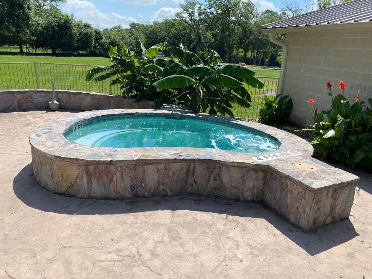 Un jacuzzi con revestimiento de piedra, rodeado por una terraza de hormigón, con un campo de césped y árboles al fondo.