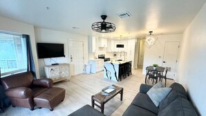 A modern living room with a sofa, armchair, and a coffee table. There is a kitchen area with a dining table and chairs, and a TV mounted on the wall. - Hill Country Hideout | Sleeps 10 | WiFi | 2 Patios (New Braunfels)