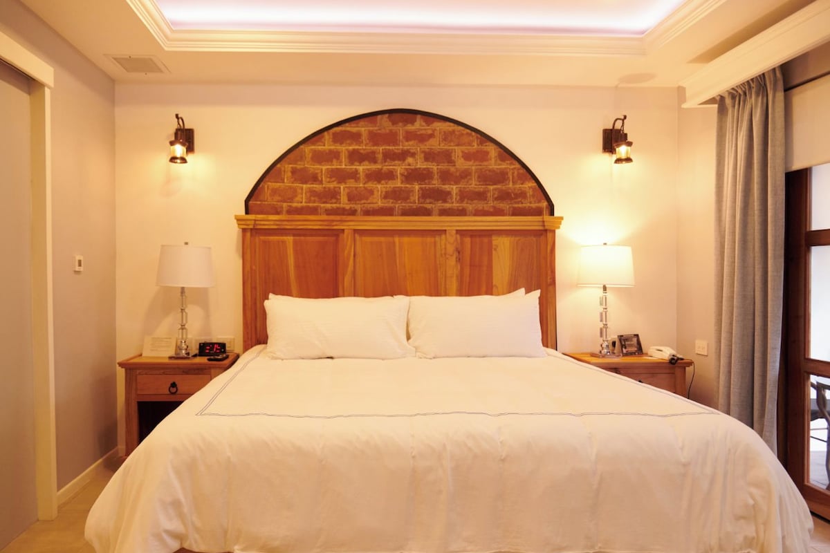 Una cama grande con tablero de madera, dos mesitas de noche con lámparas, y un arco de pared de ladrillo sobre la cama.