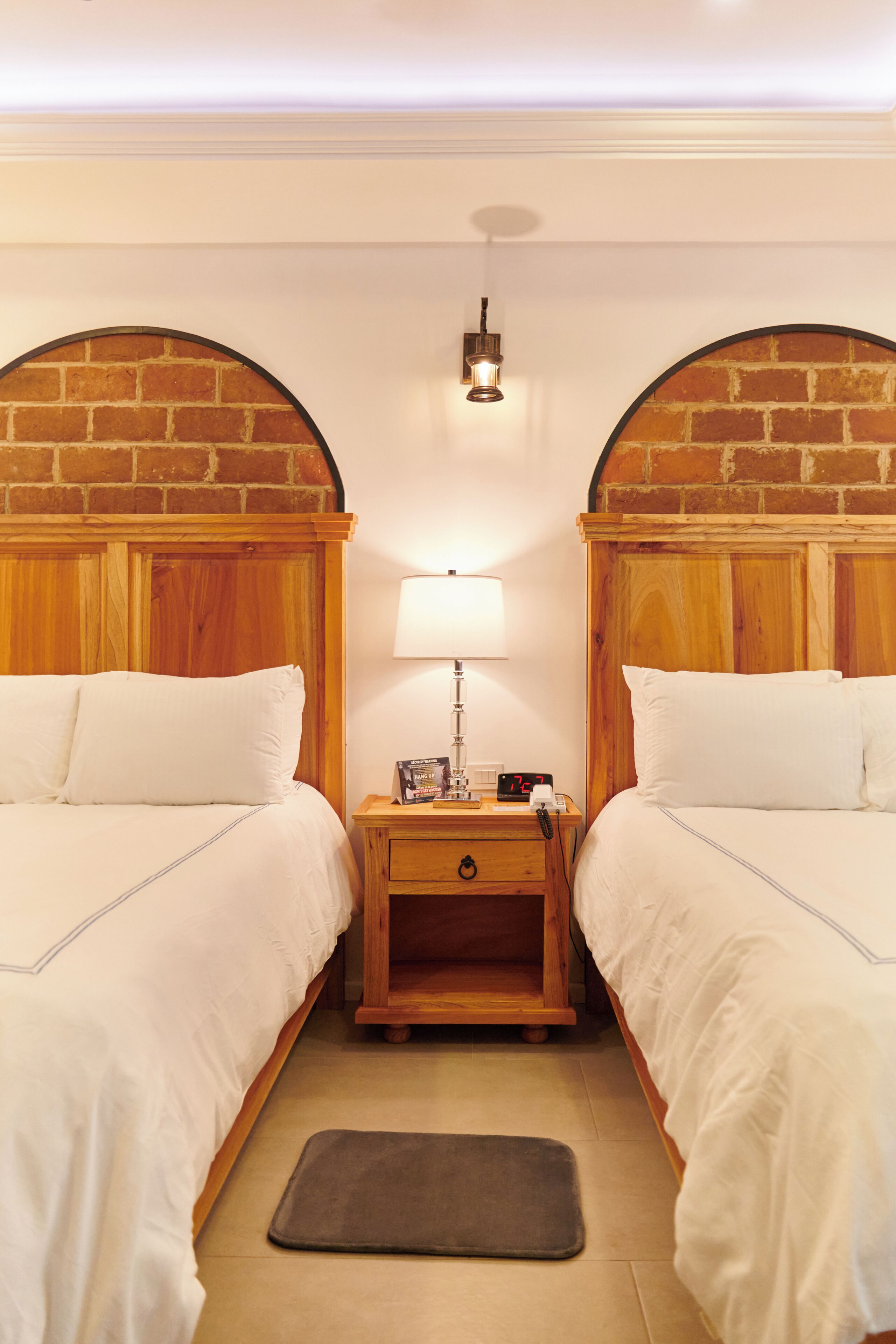 Une chambre avec deux lits, une table de chevet avec une lampe et un mur en briques.