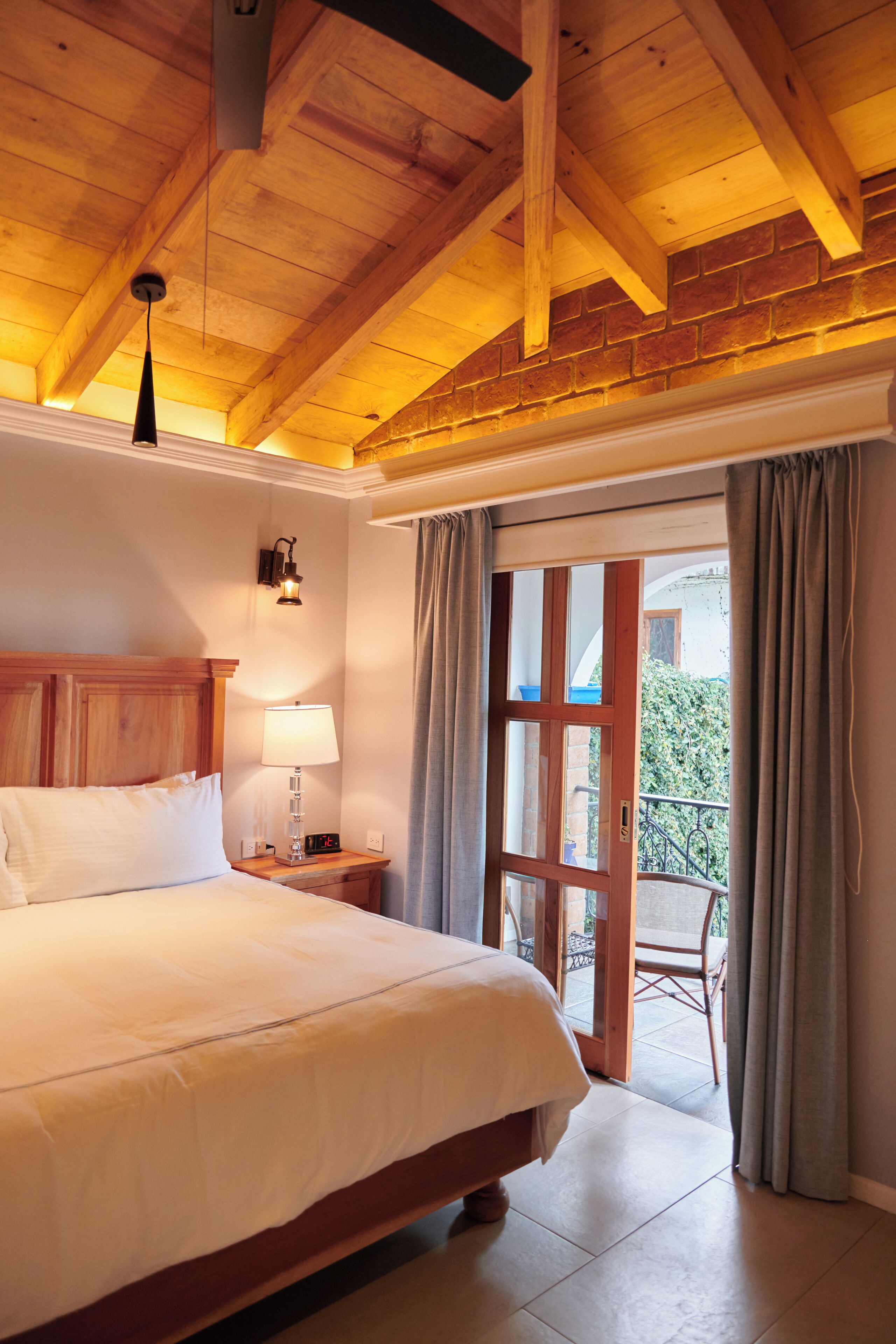 A bedroom with a wooden ceiling, a bed, a nightstand with a lamp, a window with curtains, and a view of the outside.