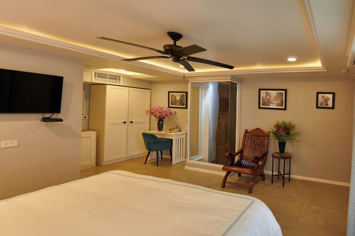 A hotel room with a bed, a desk with a chair, a ceiling fan, and a TV mounted on the wall.