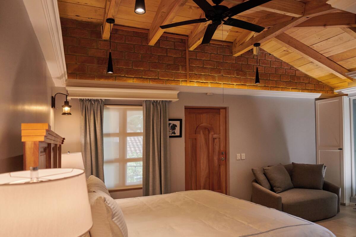 A bedroom with a large bed, a wooden headboard, a bedside table with a lamp, a remote control, and a window with curtains.