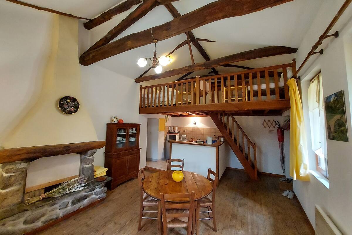 Un intérieur rustique avec une table à manger en bois, une cheminée en pierre, un escalier en bois et une chambre à coucher située en mezzanine.
