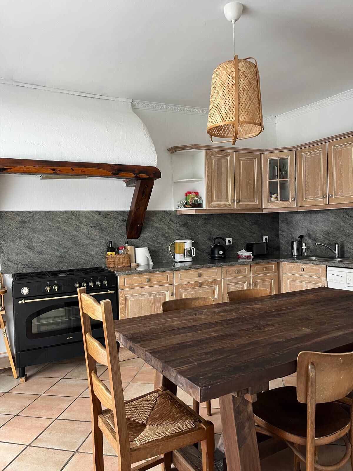 Een keuken met houten kasten, een zwart fornuis, een houten eettafel en een geweven lamp.