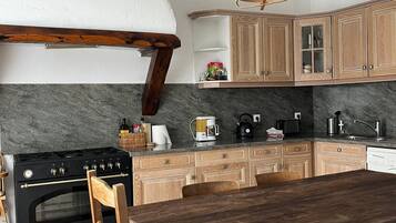 Een keuken met houten kasten, een zwart fornuis, een houten eettafel en een geweven lamp.