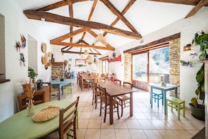 Uma sala de jantar ampla com mesas e cadeiras de madeira, teto alto com vigas aparentes e grandes janelas com vista para o exterior.