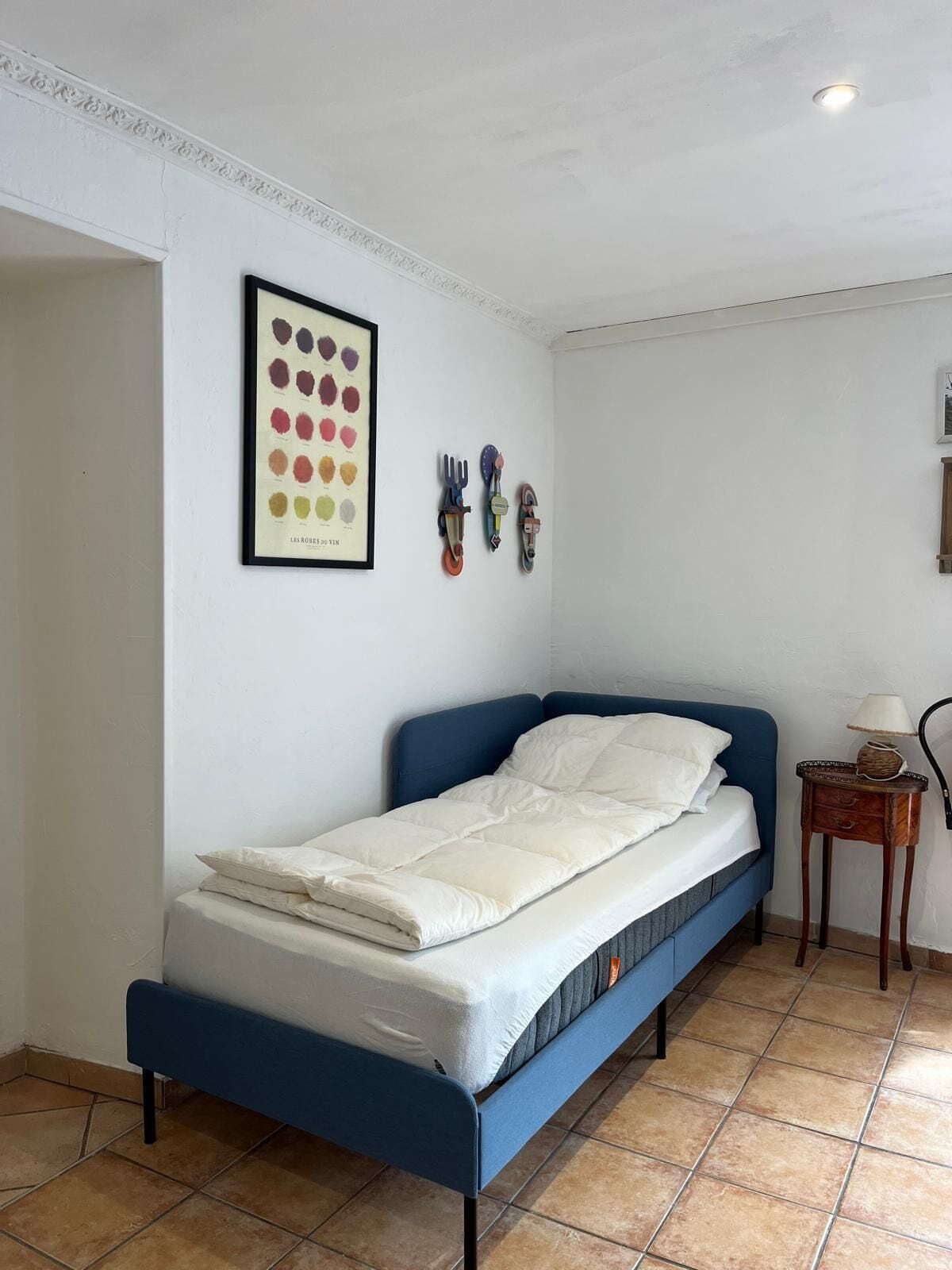 Une chambre avec un lit simple, une tête de lit bleue, du linge de lit blanc, une palette de couleurs encadrée accrochée au mur et une petite table de chevet en bois avec une lampe.