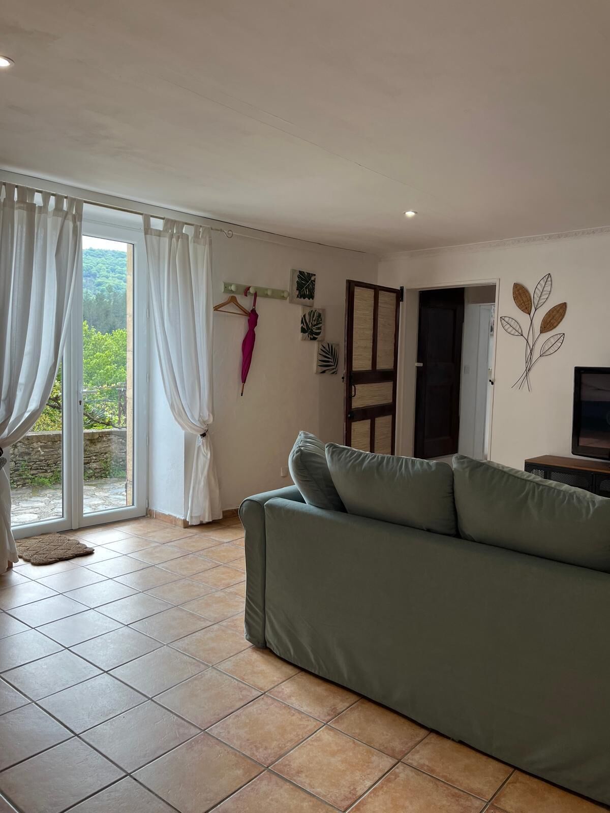 Un salon avec un canapé vert, une porte-fenêtre coulissante donnant sur un balcon et un mur décoré de motifs représentant des feuilles.