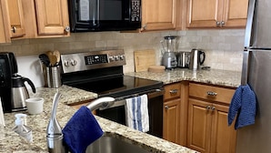 A kitchen with granite countertops, wooden cabinets, a stainless steel sink, a black microwave, a stove, and a refrigerator.