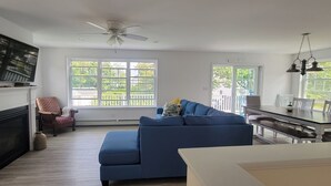 A spacious living room with a fireplace, a blue sofa, a dining table with chairs, and large windows offering a view of the outdoors.
