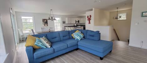 A modern living room with a blue sectional sofa, a dining area with white chairs, and a kitchen in the background.
