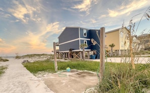 A beachfront house with a sandy path leading to it, surrounded by grass and dunes.