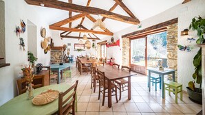 A spacious dining area with wooden tables and chairs, a high ceiling with exposed beams, and large windows offering a view of the outdoors. - Les Copains à Bord - Gîte D'étape (Cans et Cévennes)