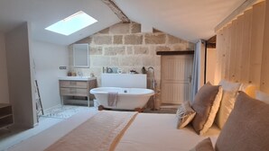 Un dormitorio con una cama grande, tragaluces, pared de piedra, puerta de madera y baño con bañera.