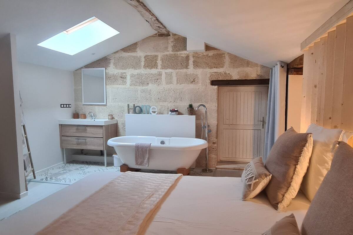 A bedroom with a large bed, a skylight, a stone wall, a wooden door, and a bathroom with a bathtub.