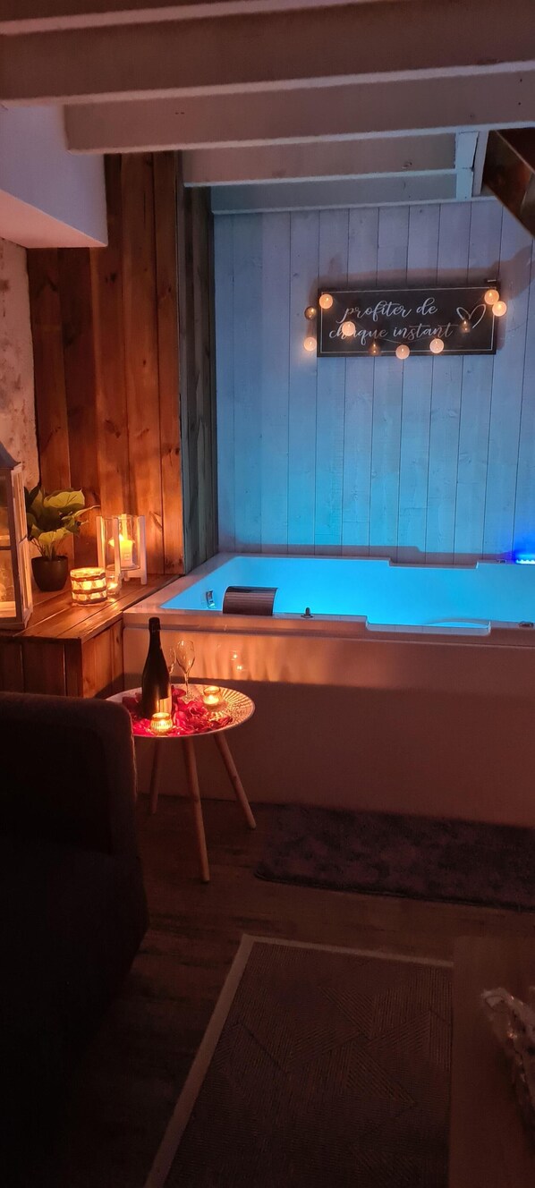 A cozy room with a wooden wall, a small table with candles, and a sign above a bathtub. - Lariviere (Asques)