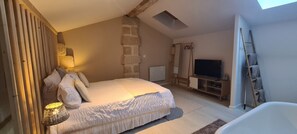 A bedroom with a large bed, a television, a stone wall, and a skylight. - Lariviere (Asques)
