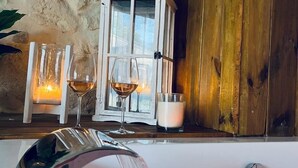 Una habitación con pared de piedra, puerta de madera, armario blanco y una mesa con dos copas de vino y una vela.