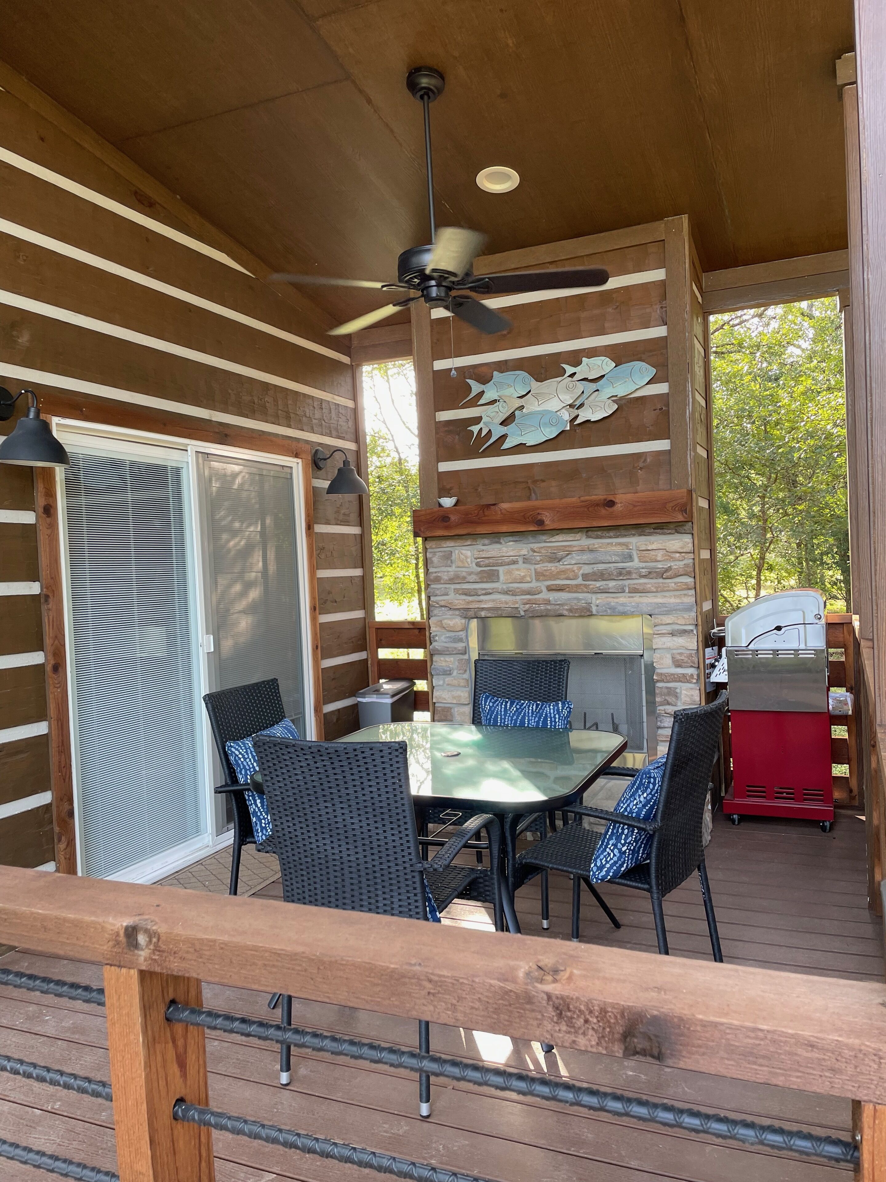 An outdoor patio with a table and chairs, a ceiling fan, a stone fireplace, and a grill.