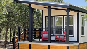 A covered outdoor seating area with a wooden deck and a metal railing, featuring two red chairs and a table. - Outcast Landing - A Fisherman's Paradise (Cedar Creek)