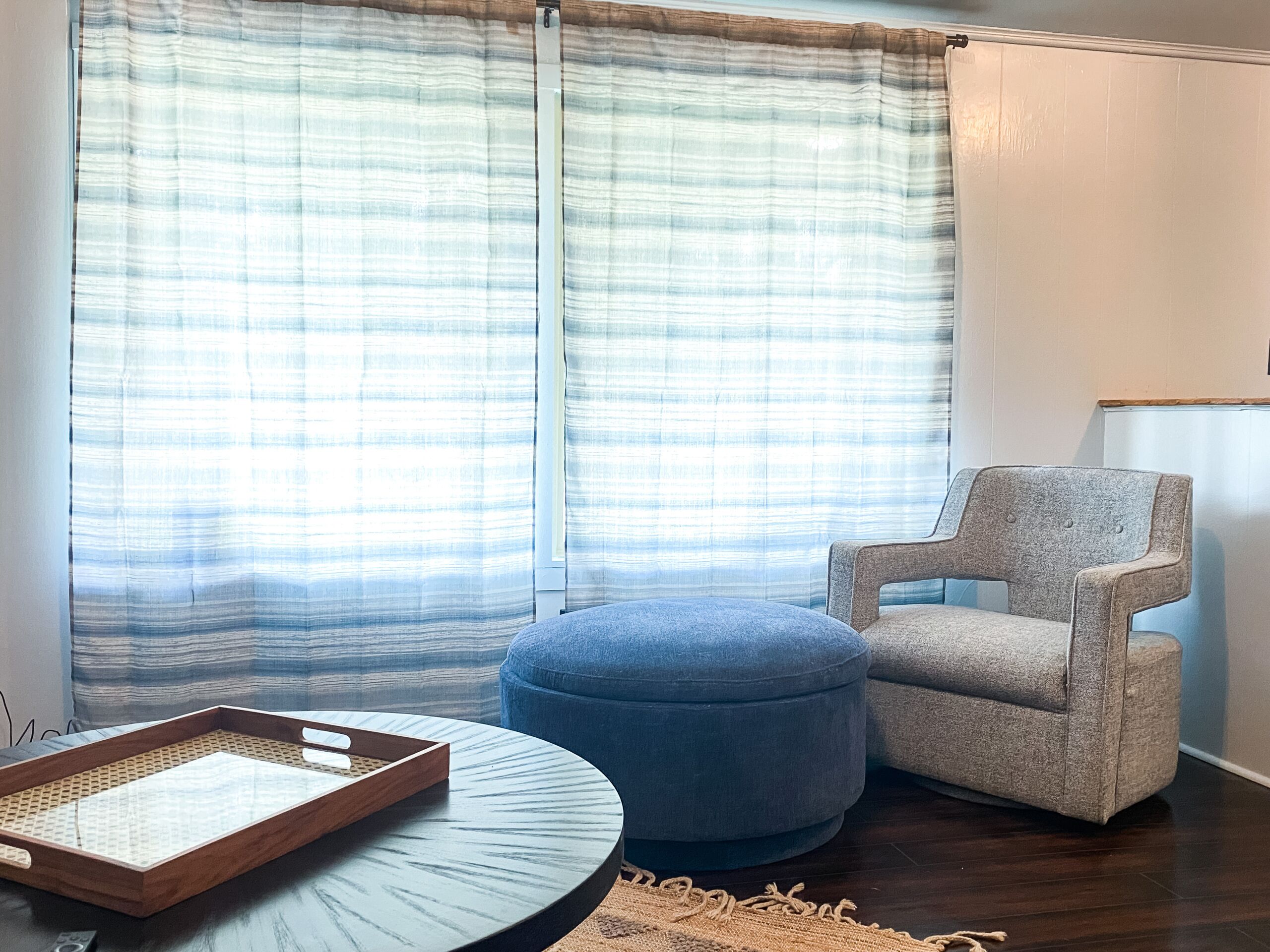 Una habitación con una mesa redonda, un pouf azul, un sillón gris y una bandeja de madera.