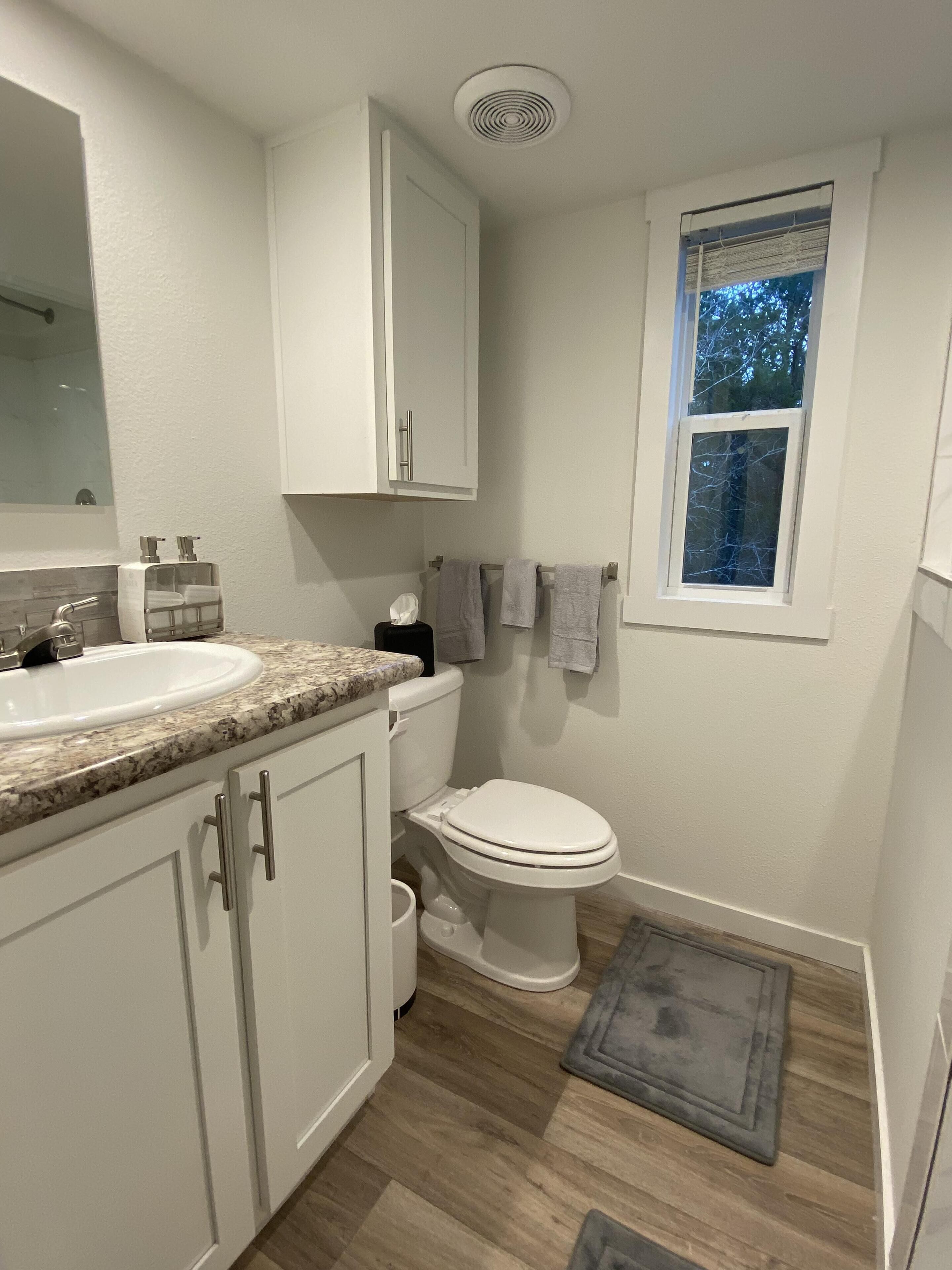 Een badkamer met een witte wastafel, toilet en kast, een raam met uitzicht op bomen, en een houten vloer.